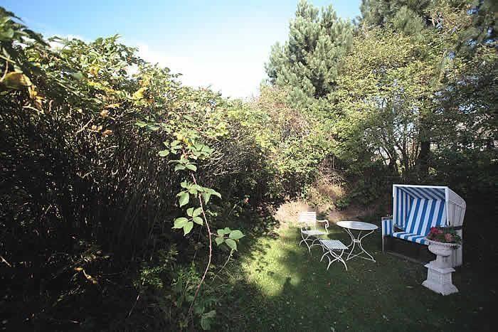 Garten mit weißem Tisch, Stühlen und Strandkorb. Umgeben von grünen Sträuchern und Bäumen.