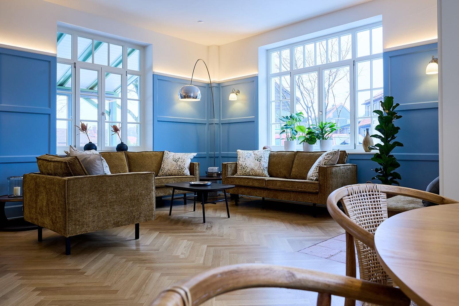 Wohnzimmer mit blauen Wänden, Holzboden und zwei Sofas. Große Fenster und Pflanzen sorgen für viel Licht.