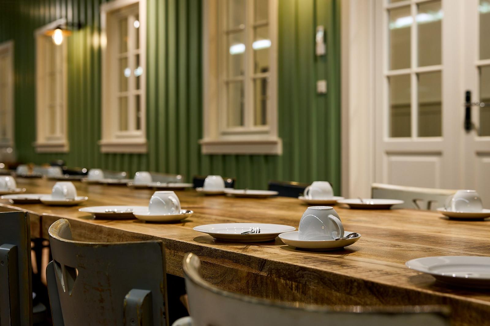 Langtisch mit Tassen und Tellern in einem Raum mit grünem Holzpaneelen und Fenstern.
