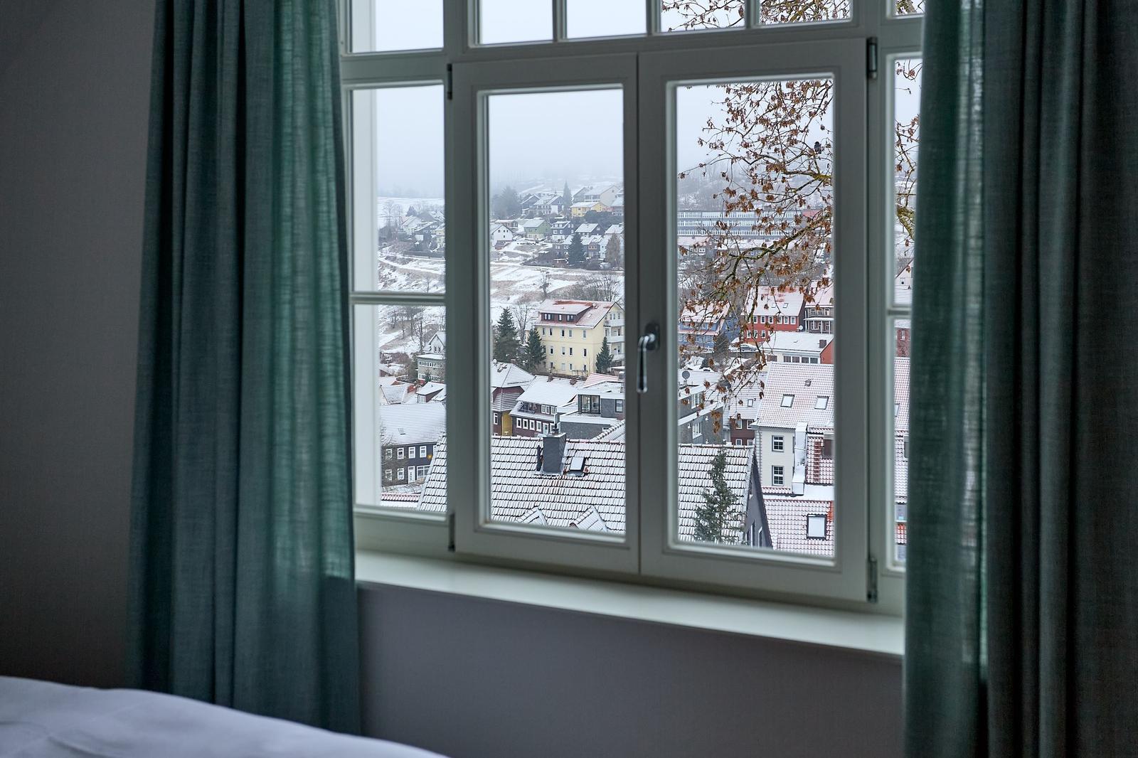 Durch das Fenster blickt man auf eine schneebedeckte Stadtlandschaft mit Dächern und Bäumen.