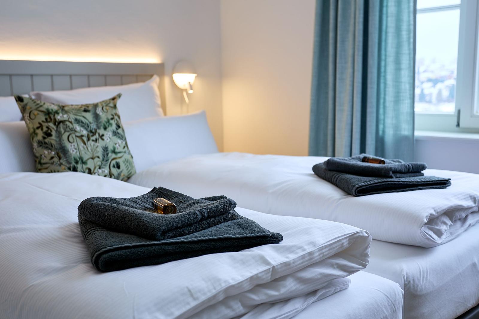 Zwei Betten mit weißen Laken und dunklen Handtüchern. Fenster mit blauen Vorhängen.
