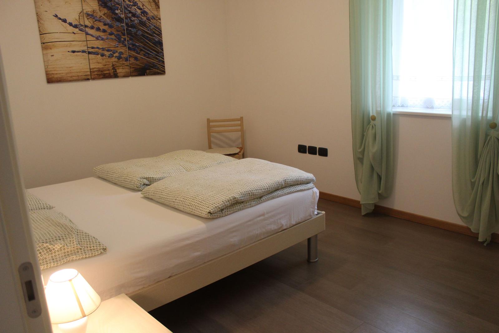 Schlafzimmer mit Bett, Wandbild, Stuhl und Fenster mit Vorhängen.