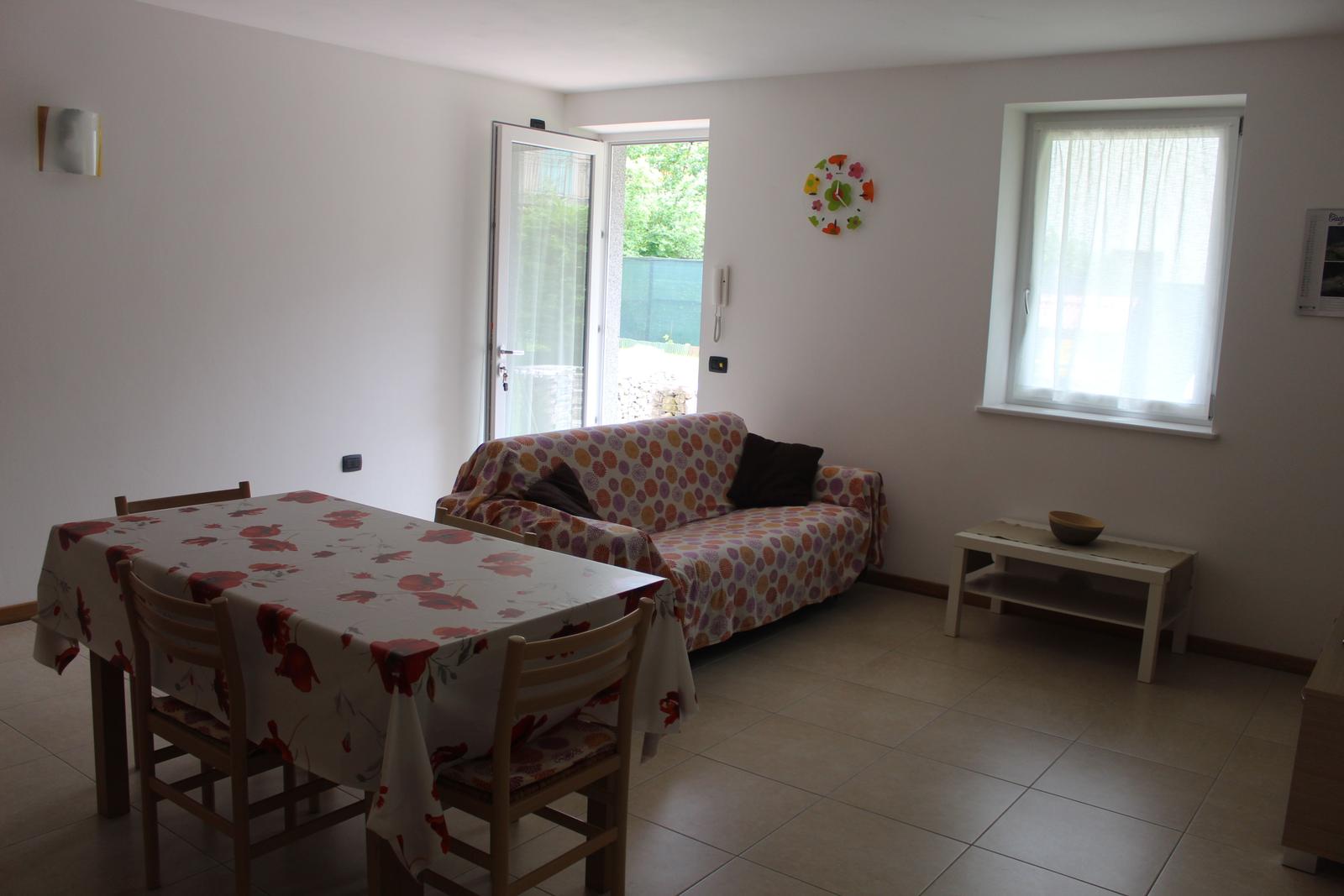 Wohn- und Essbereich mit Tisch, Stühlen und Sofa. Fenster und Glastür mit Blick nach draußen.