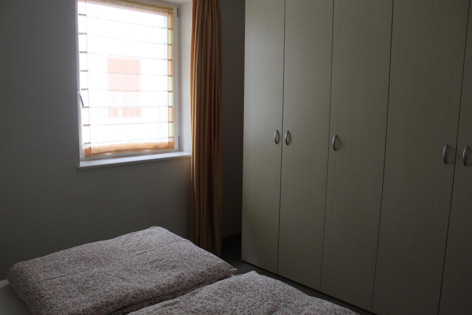 Schlafzimmer mit Bett, Kleiderschrank und Fenster mit Jalousie und Vorhang.