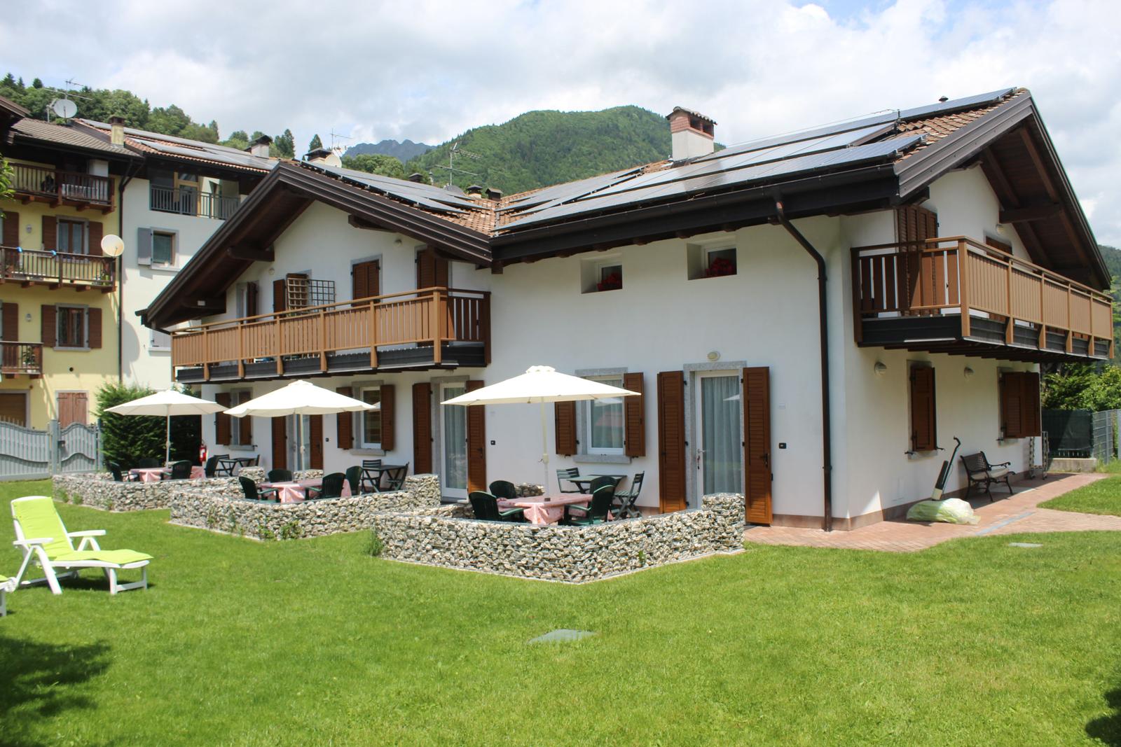Ein zweistöckiges Haus mit Balkonen, Terrasse und Garten. Im Hintergrund sind Berge zu sehen.