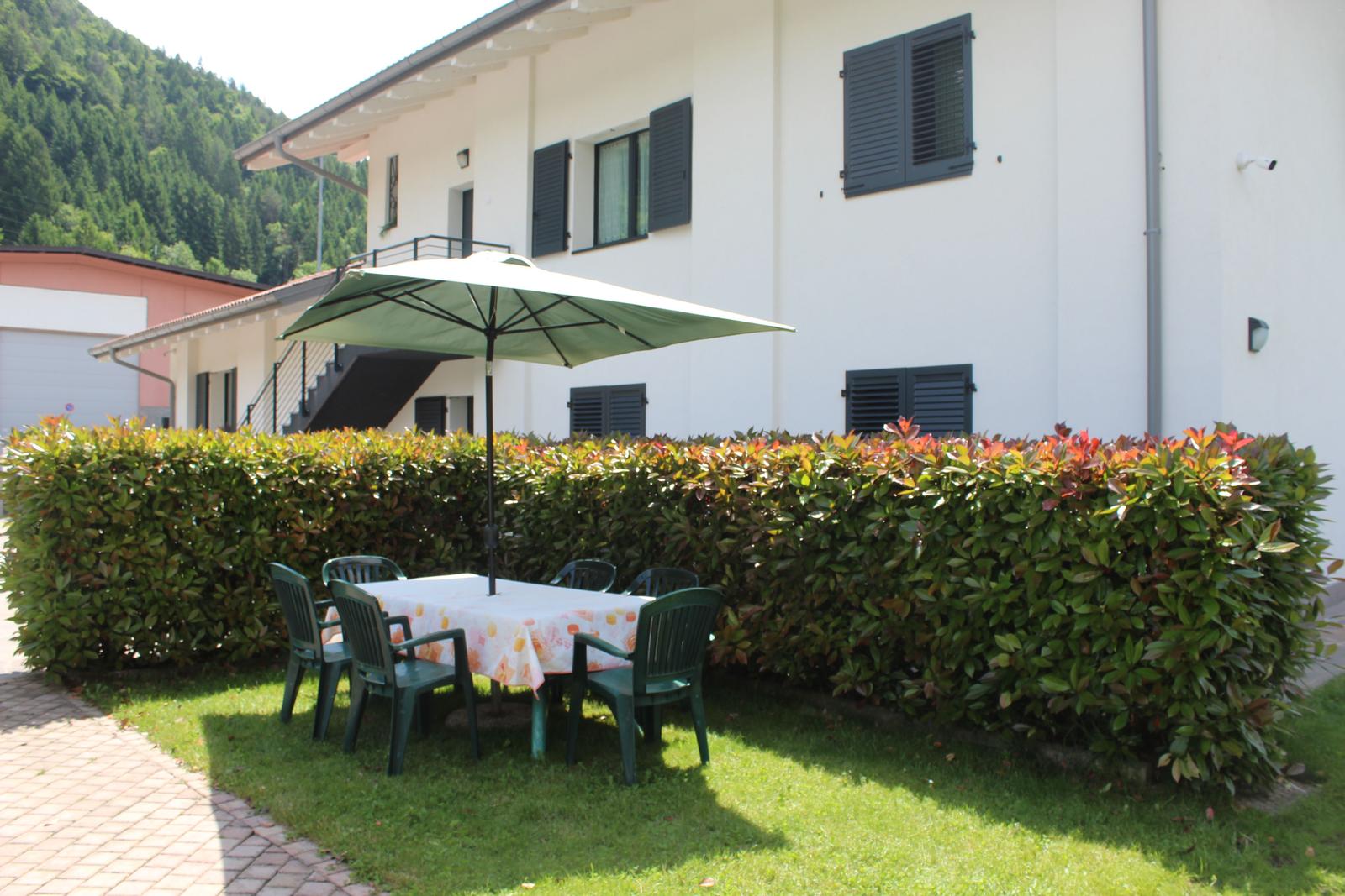 Terrasse mit Tisch, Stühlen und Sonnenschirm vor weißem Haus mit Hecke.