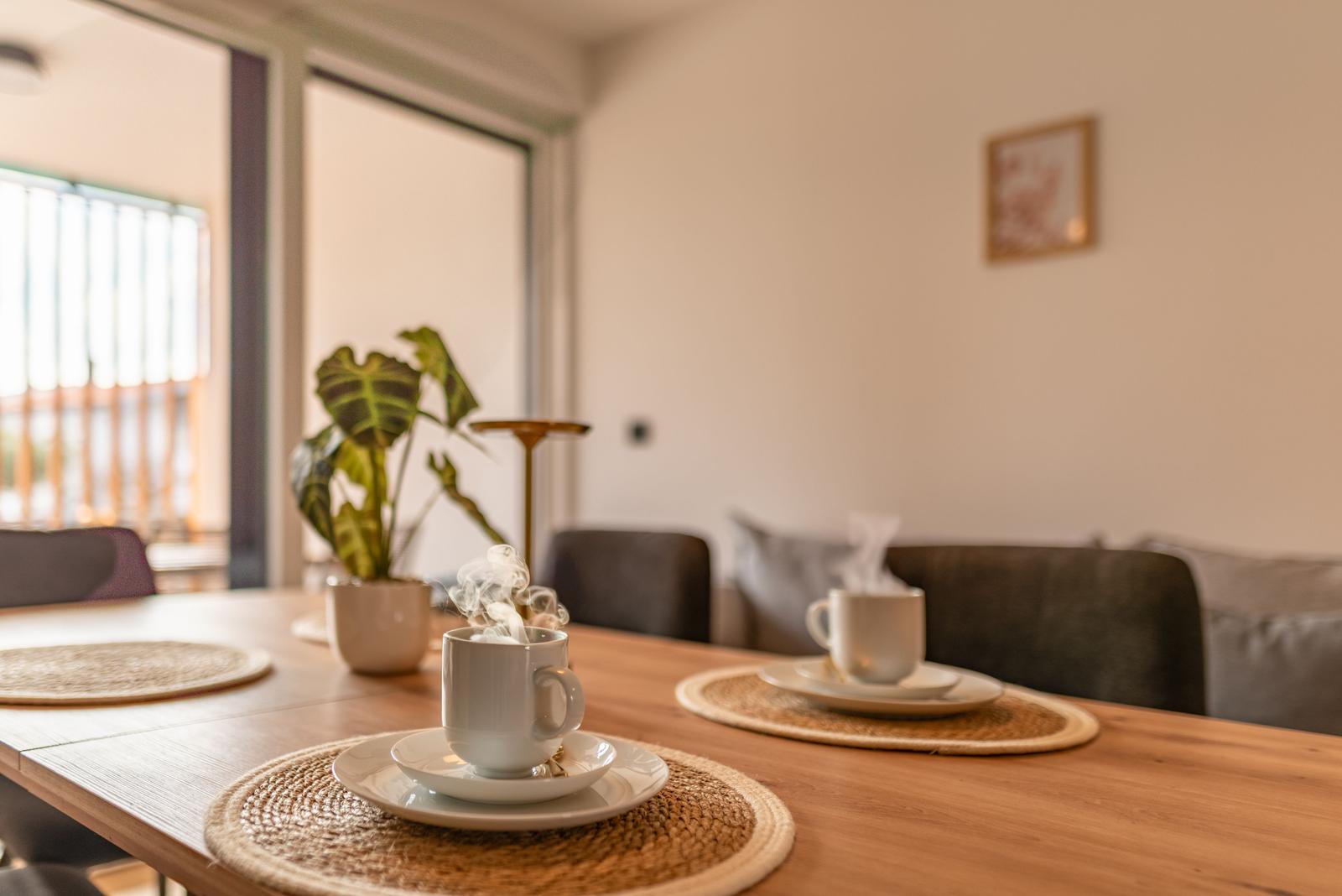 Dining table with steaming cups, plant, and seating in the background.