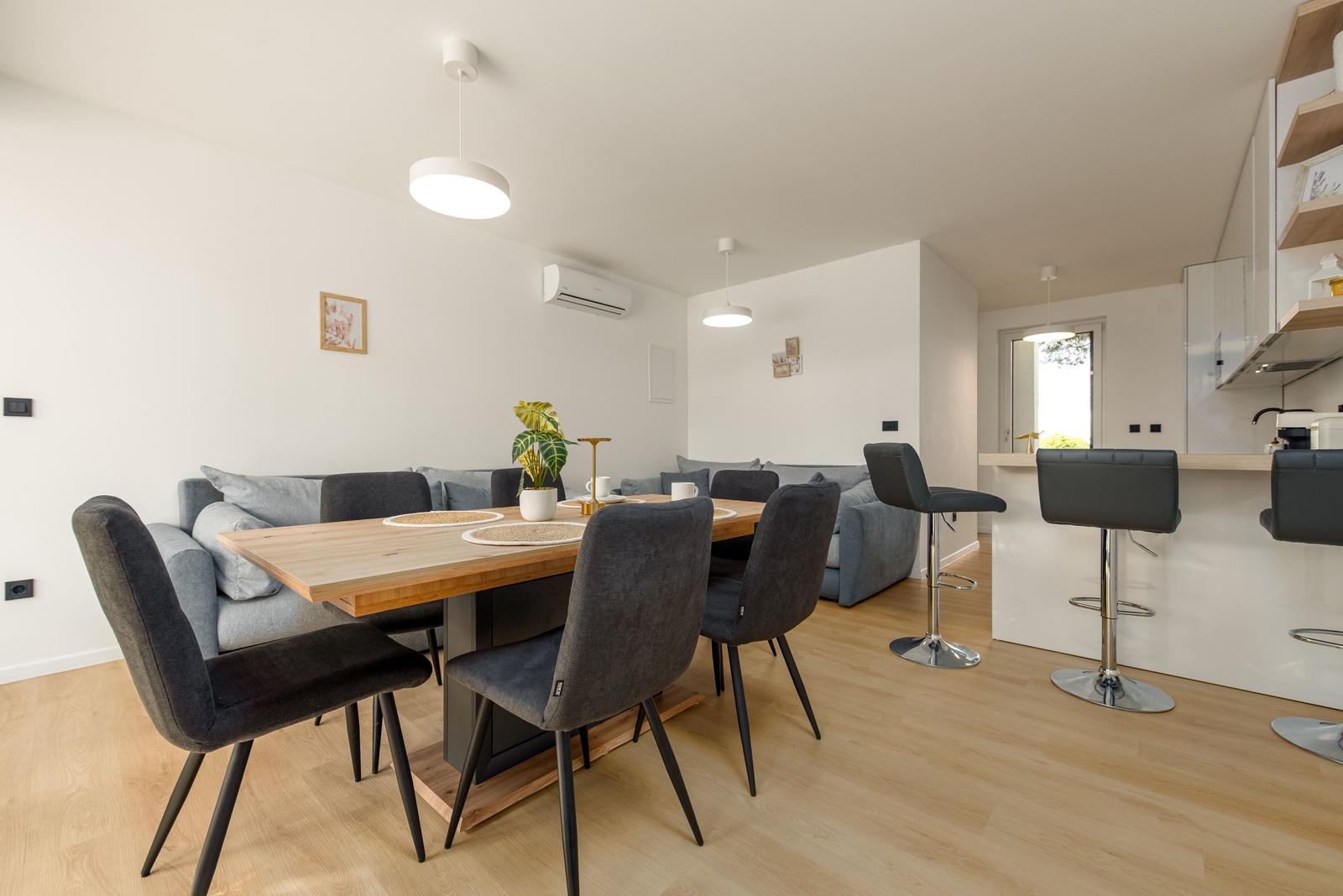 Dining area with wooden table and chairs, sofa, and kitchen counter in background.