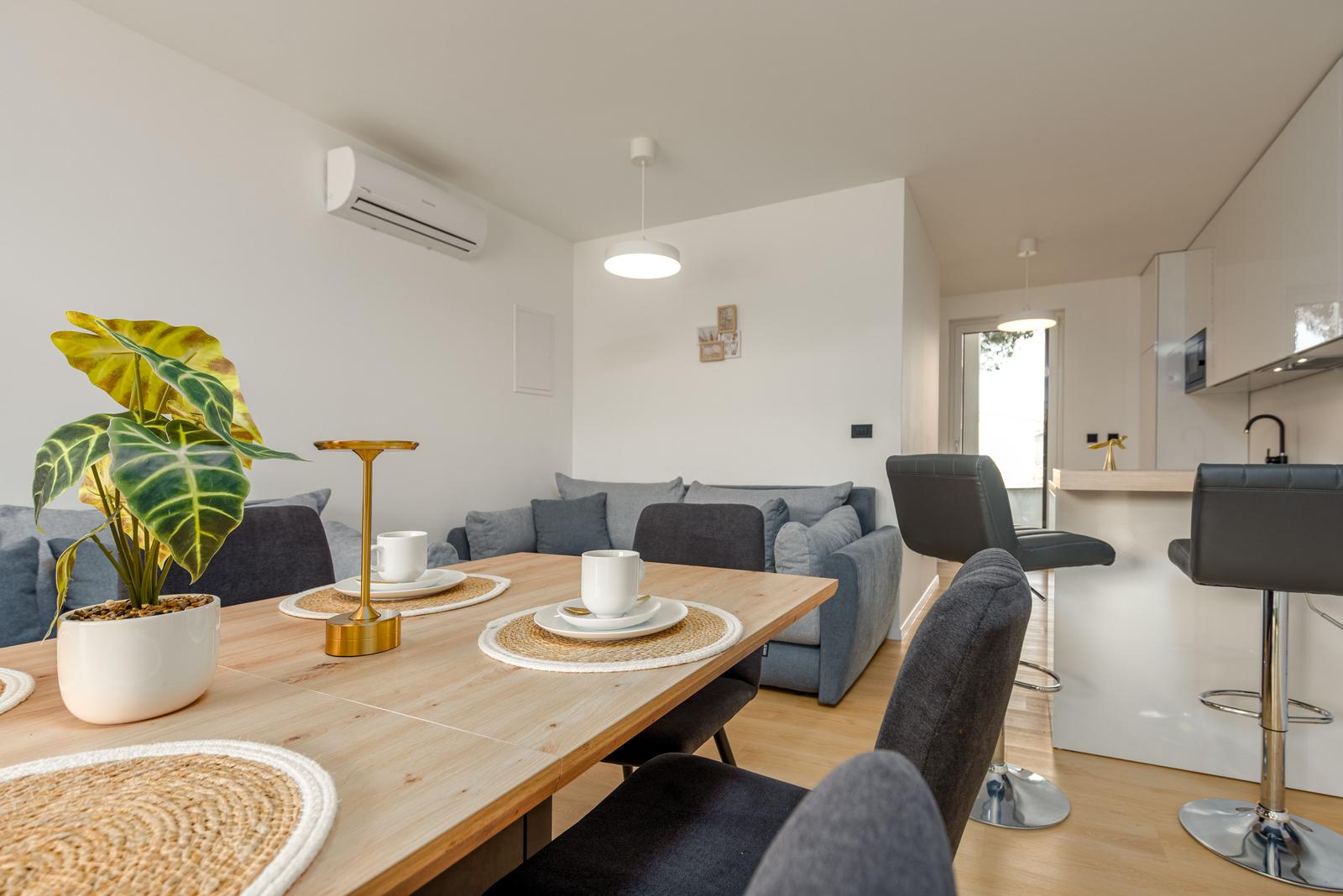 Dining area with table, chairs, and plant. Sofa and kitchen area in background.