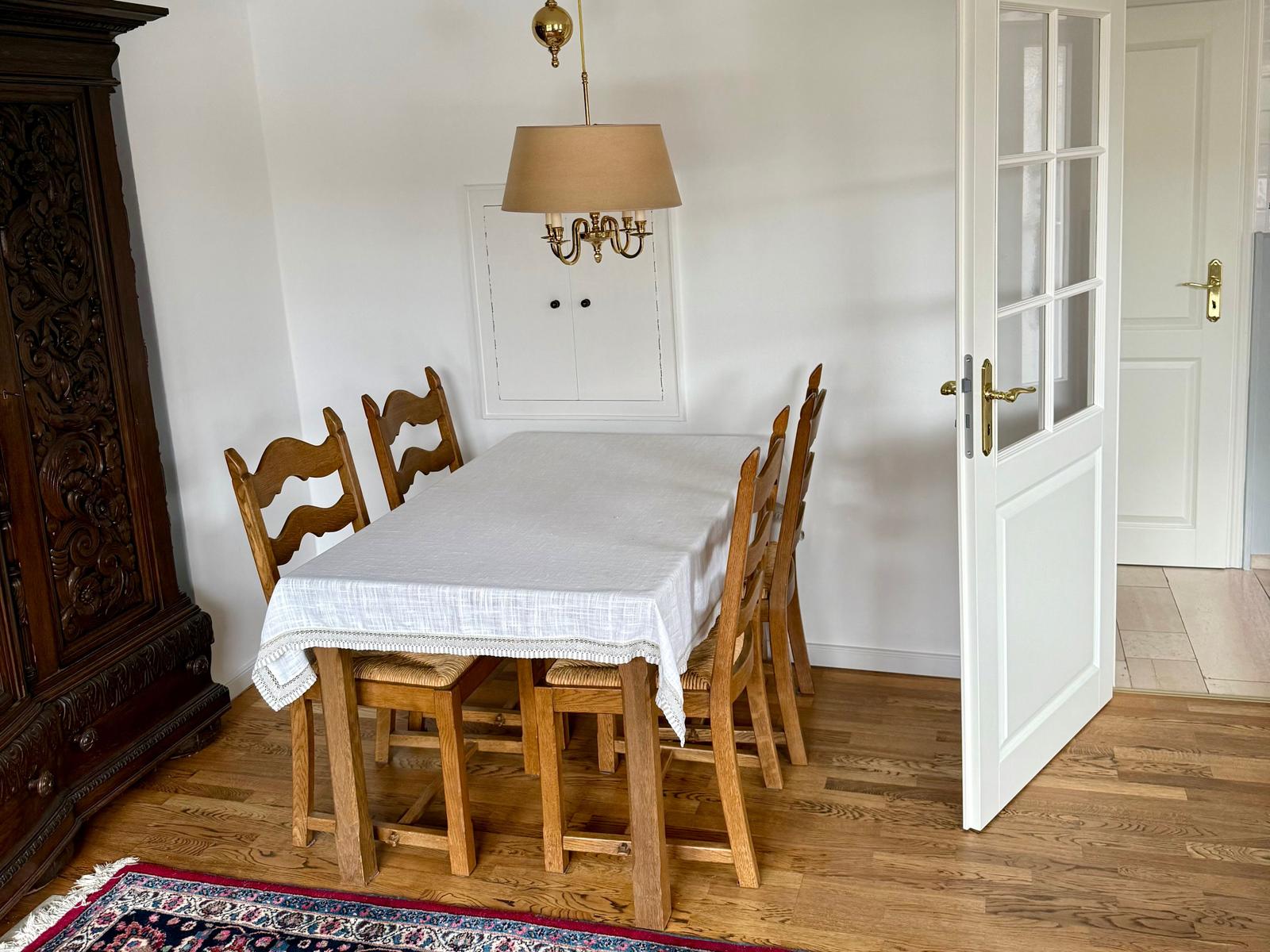 Esszimmer mit Holztisch, Stühlen und Tischdecke. Hintergrund: Tür und Wandgarderobe.