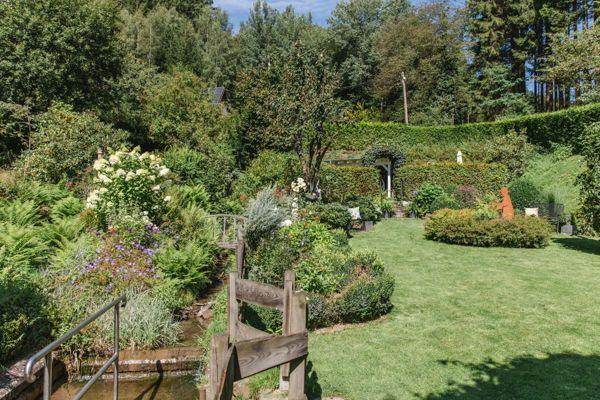 Garten mit grünem Rasen, Blumenbeeten und Holzbrücke über einen kleinen Teich.