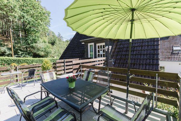 Terrasse mit Tisch, Stühlen und grünem Sonnenschirm. Hintergrund: Holzgeländer und Haus.