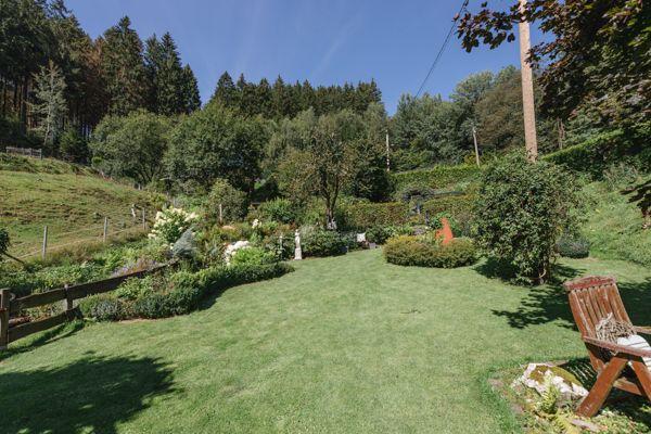 Großer grüner Garten mit Holzstuhl und Blumenbeete. Hintergrund: Wald und Hügel.