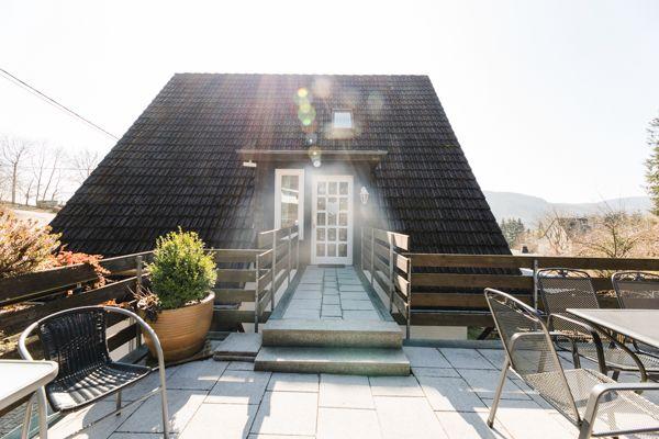 Haus mit Terrasse, Tisch, Stühlen und Pflanzen. Hintergrund: Berge und klarer Himmel.
