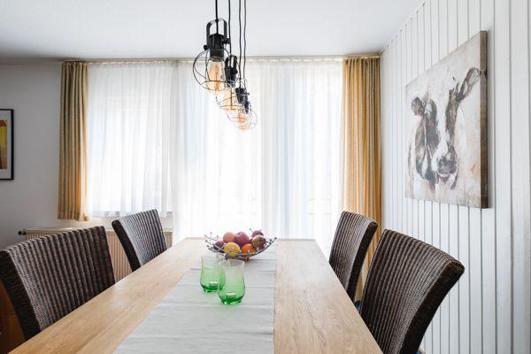 Esszimmer mit Holztisch, Stühlen und Hängelampe. Fenster mit Vorhängen und Wandbild.