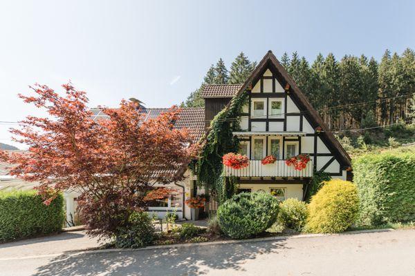 Fachwerkhaus mit Balkon, umgeben von Bäumen und Sträuchern.