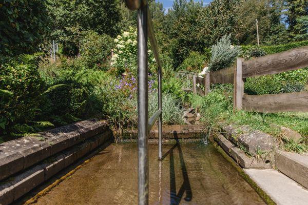 Ein kleiner Bach fließt durch einen grünen Garten mit Steinufer und Holzgeländer.