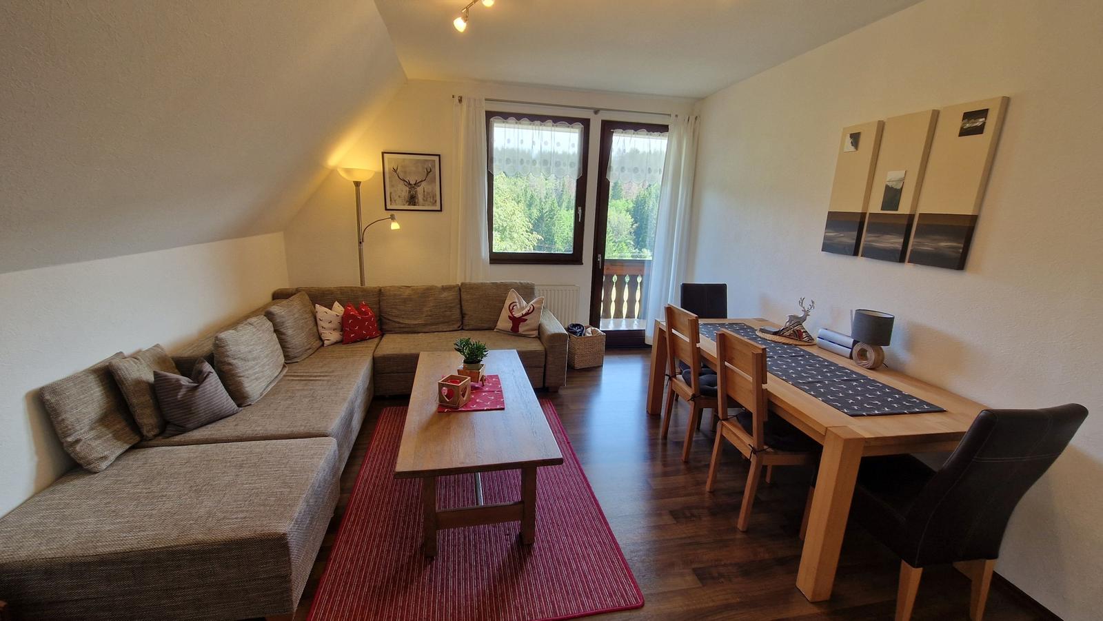 Living and dining area with sofa, table, and chairs. Window with view of balcony and forest.