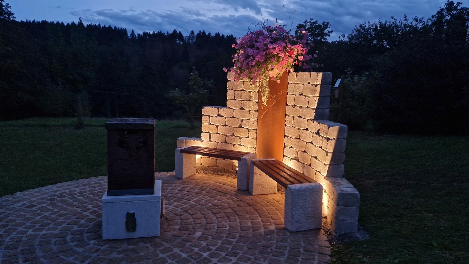 Garden area with stone bench, flower box, and lighting at dusk.