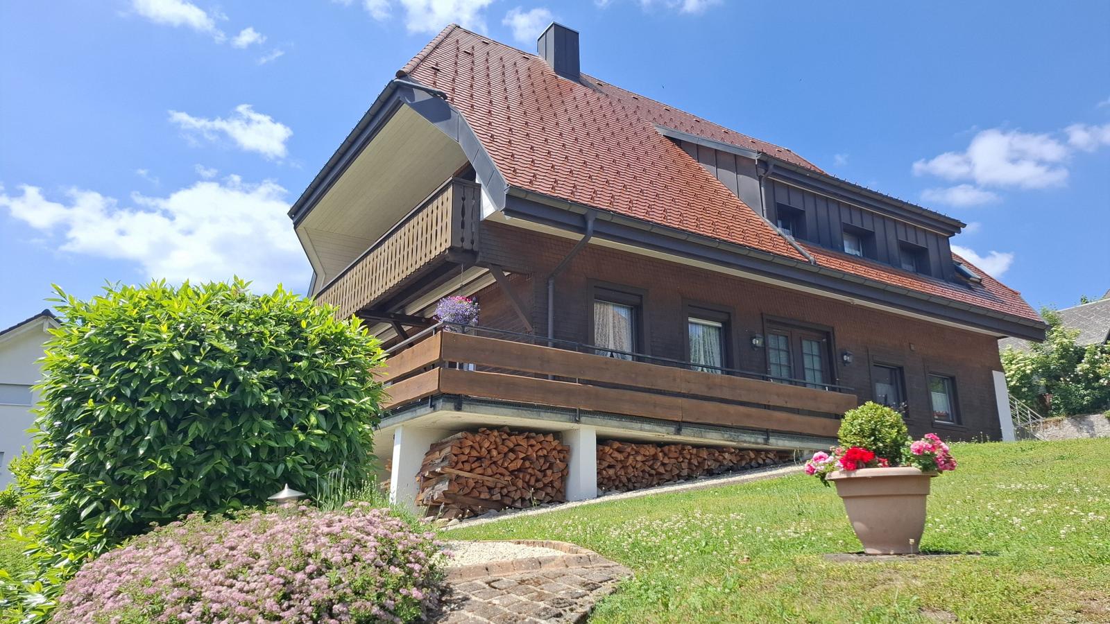 House with balcony, woodpile, and flowerbeds under blue sky.