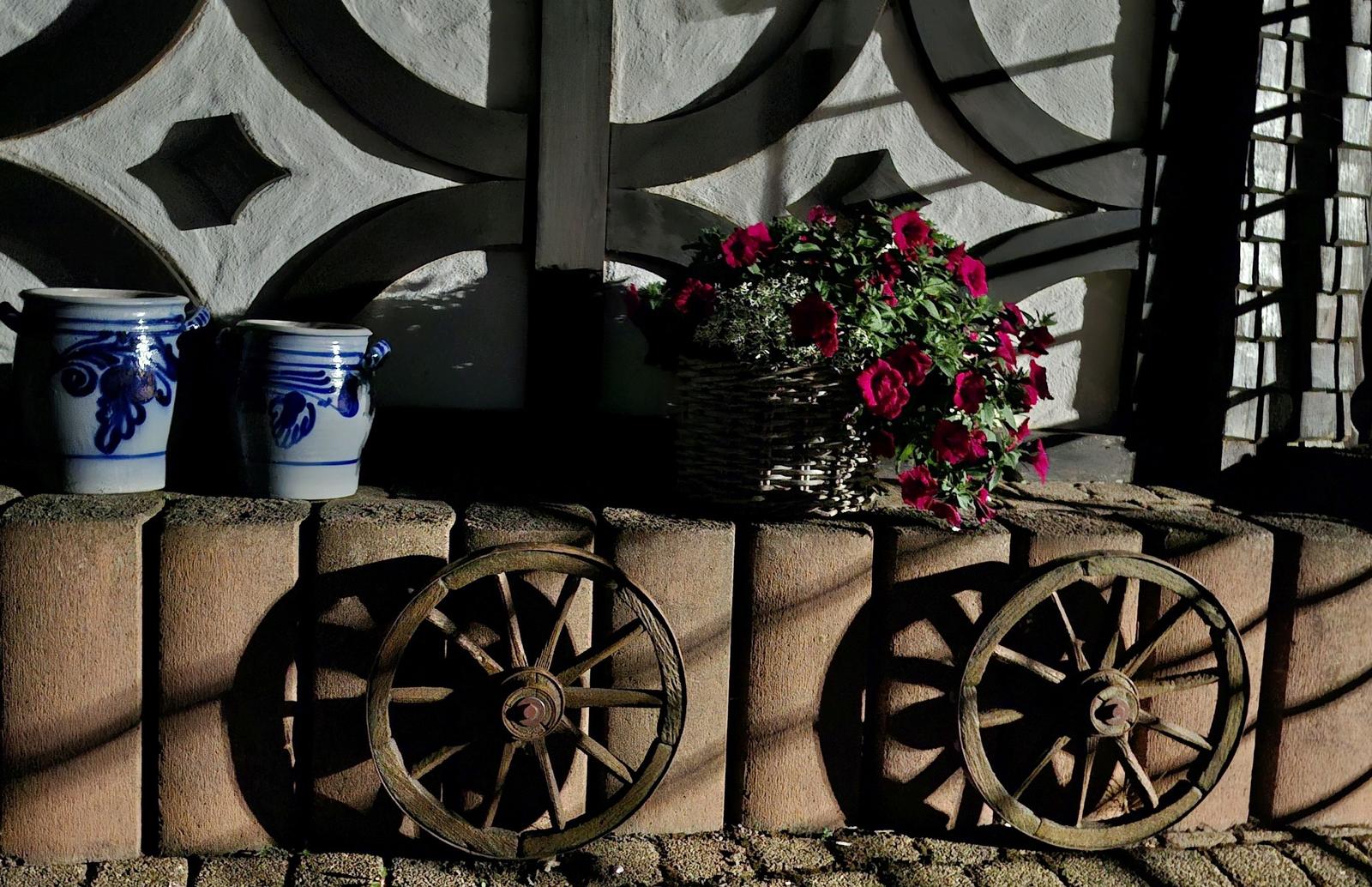 Two blue ceramic pots, a basket with red flowers, and two wooden wheels on stone wall.