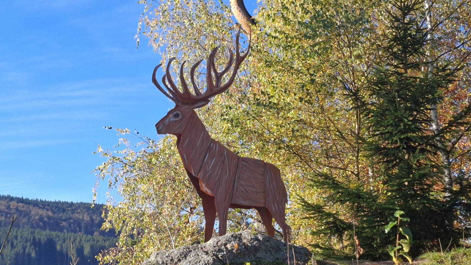 Wooden sculpture of a deer on a rock in the forest with autumn leaves.