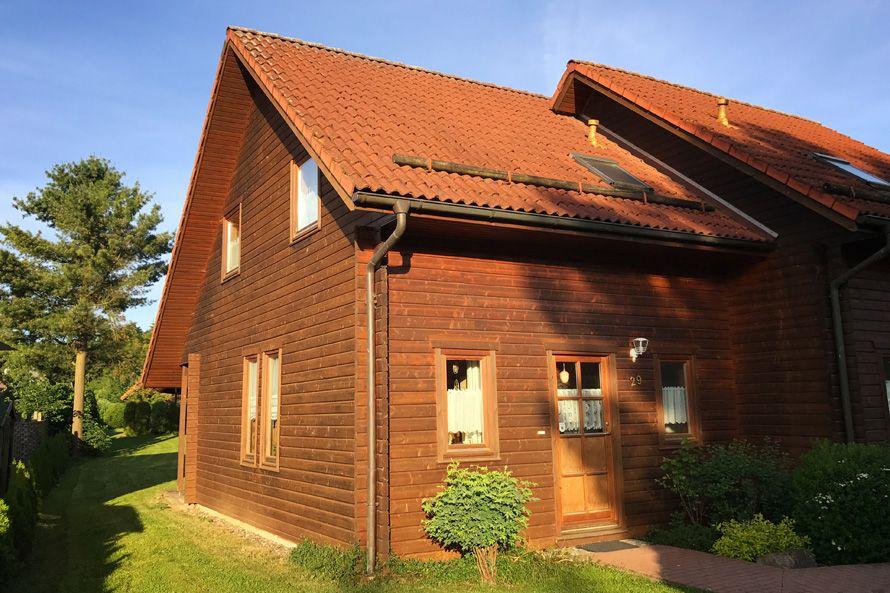 Holzhaus mit rotem Dach, Eingangstür und Fenstern, umgeben von Grünflächen und Bäumen.