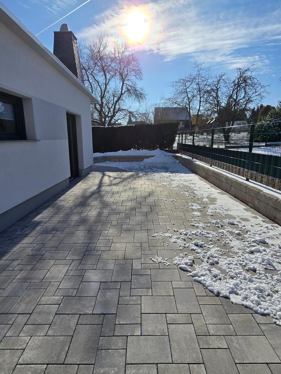 Pflasterter Weg neben Haus mit Schnee und Bäumen im Hintergrund.