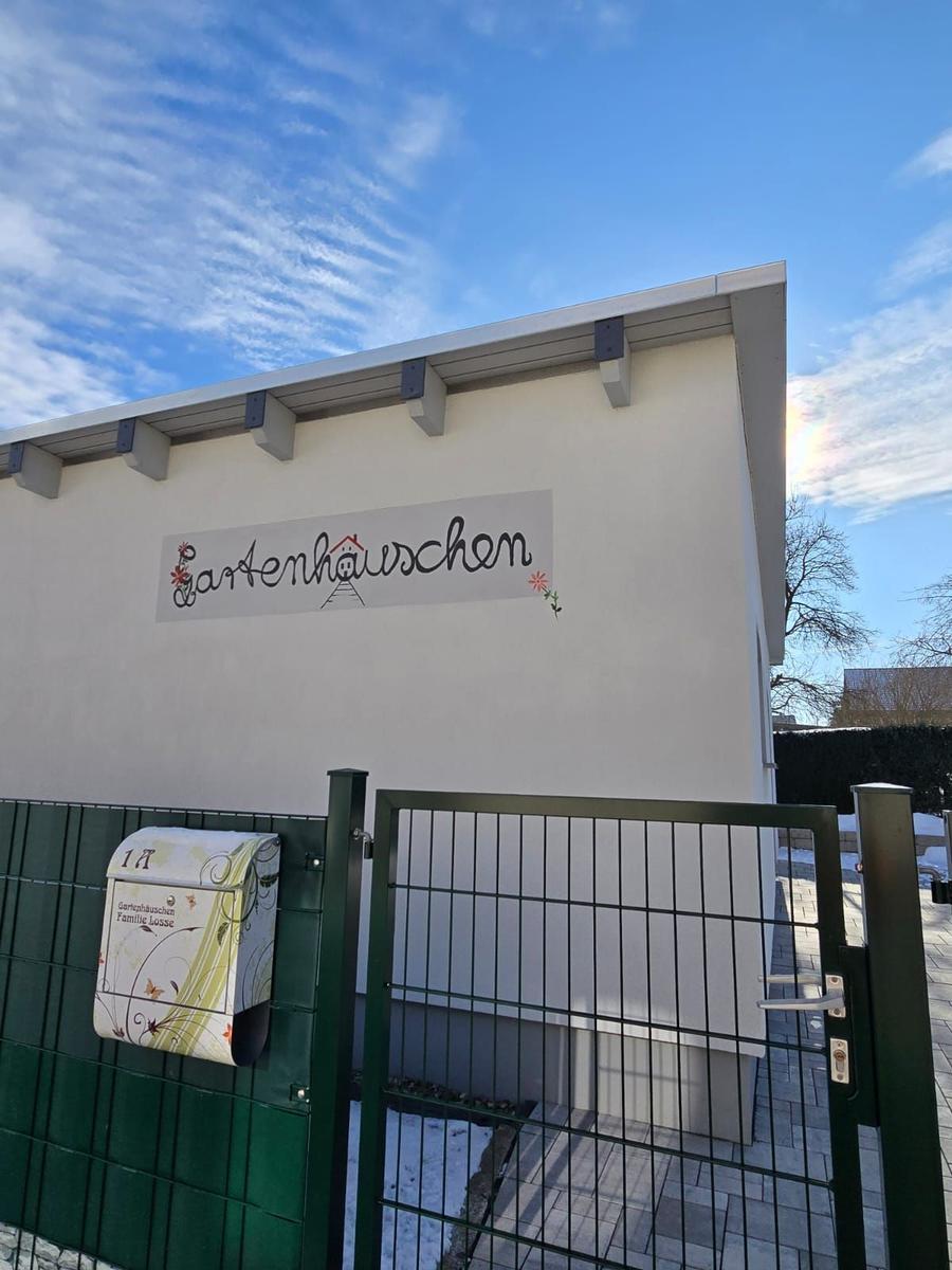 Gartenhauschen mit Briefkasten und Zaun an sonnigem Tag.