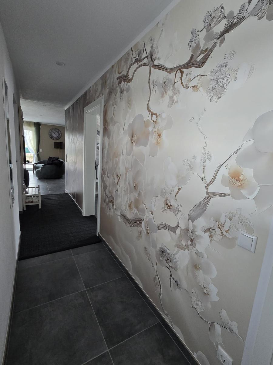 Flur mit floraler Wandtapete und dunklem Bodenbelag. Blick in Wohnbereich mit Sitzmöbeln.