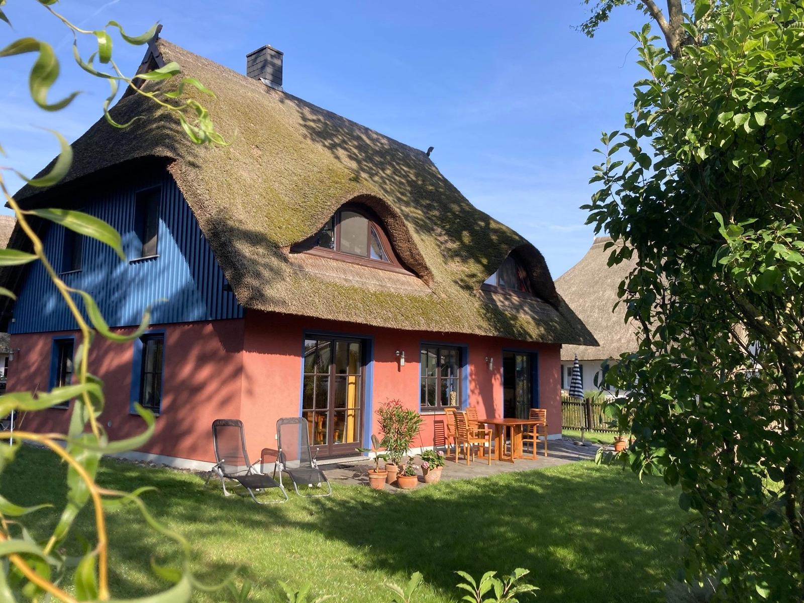 Haus mit Strohdach, Terrasse und Garten. Holztisch, Stühle und Sonnenschutz.