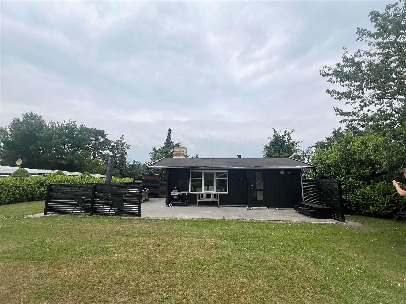 Black cabin with terrace, grill, and garden. Surrounded by trees and shrubs.
