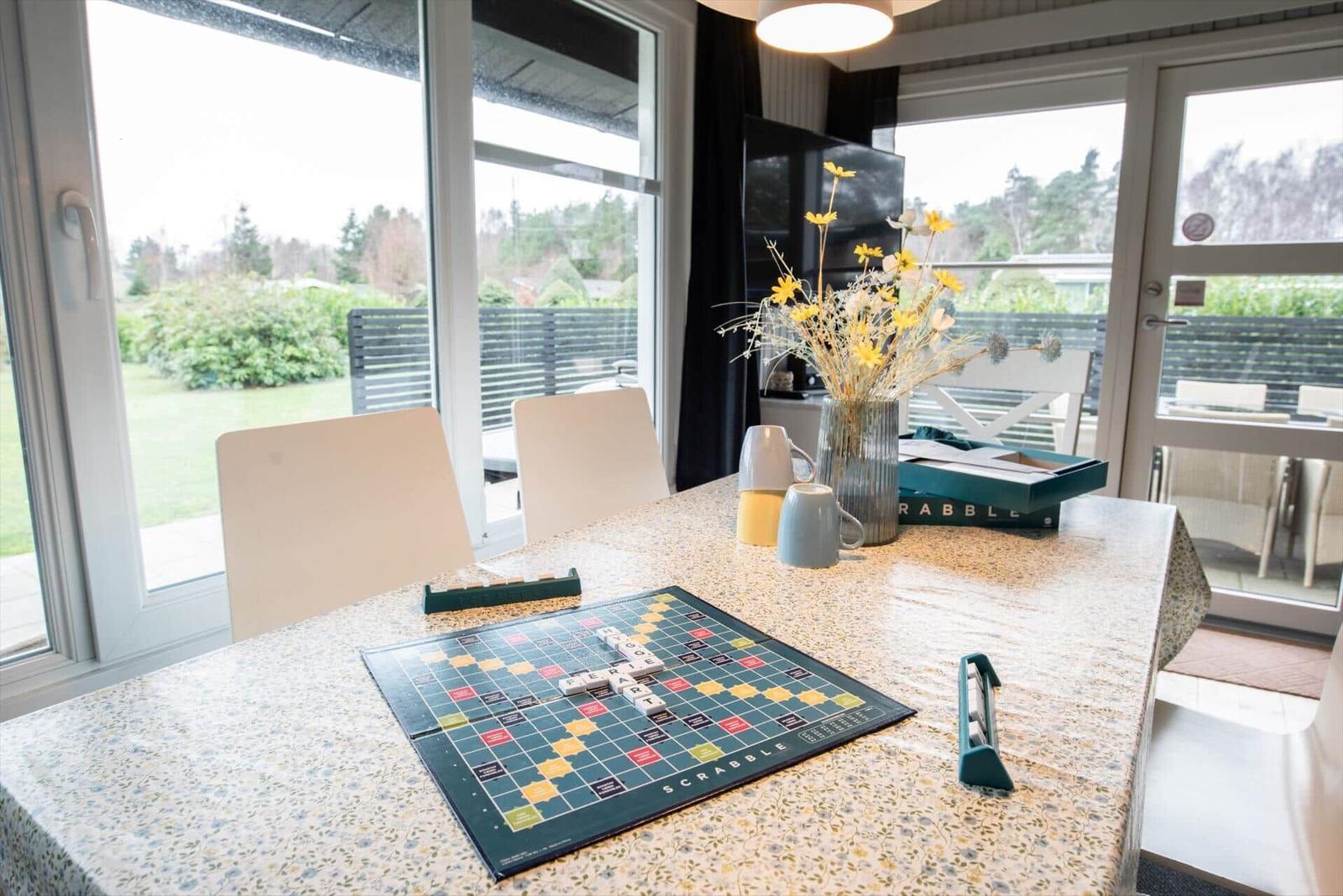 Dining table with Scrabble game, flowers and mugs by windows overlooking garden.