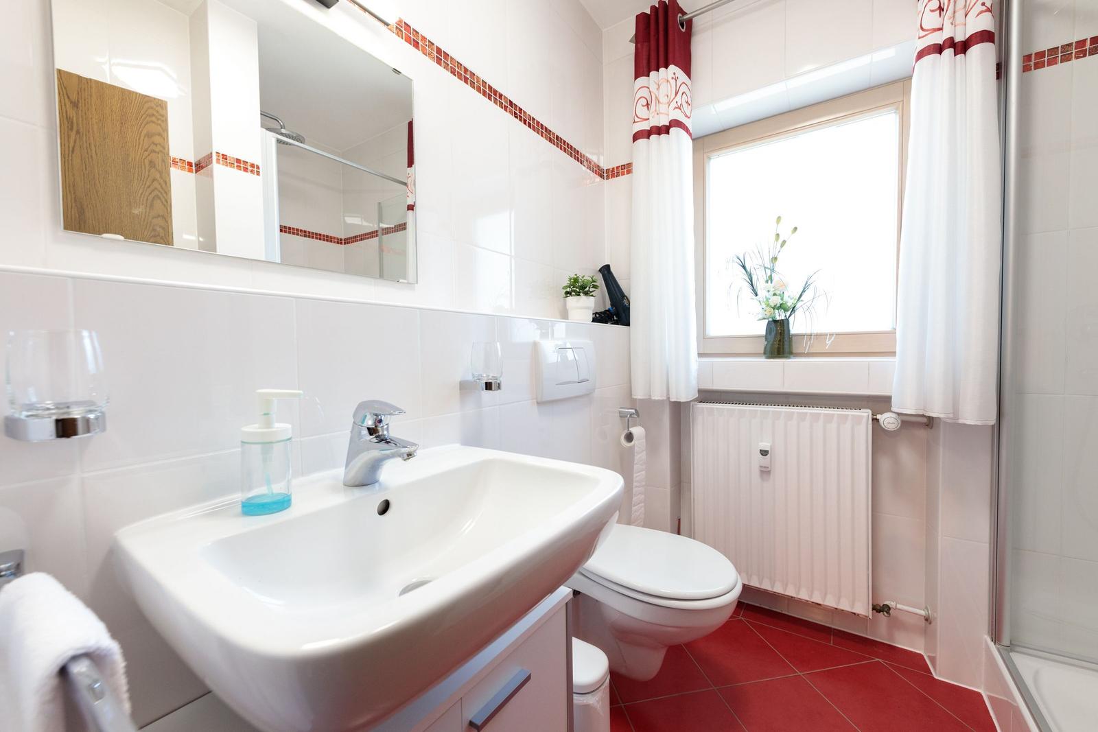 Badezimmer mit Waschbecken, Toilette und Dusche. Rote Fliesen und Fenster mit Vorhang.