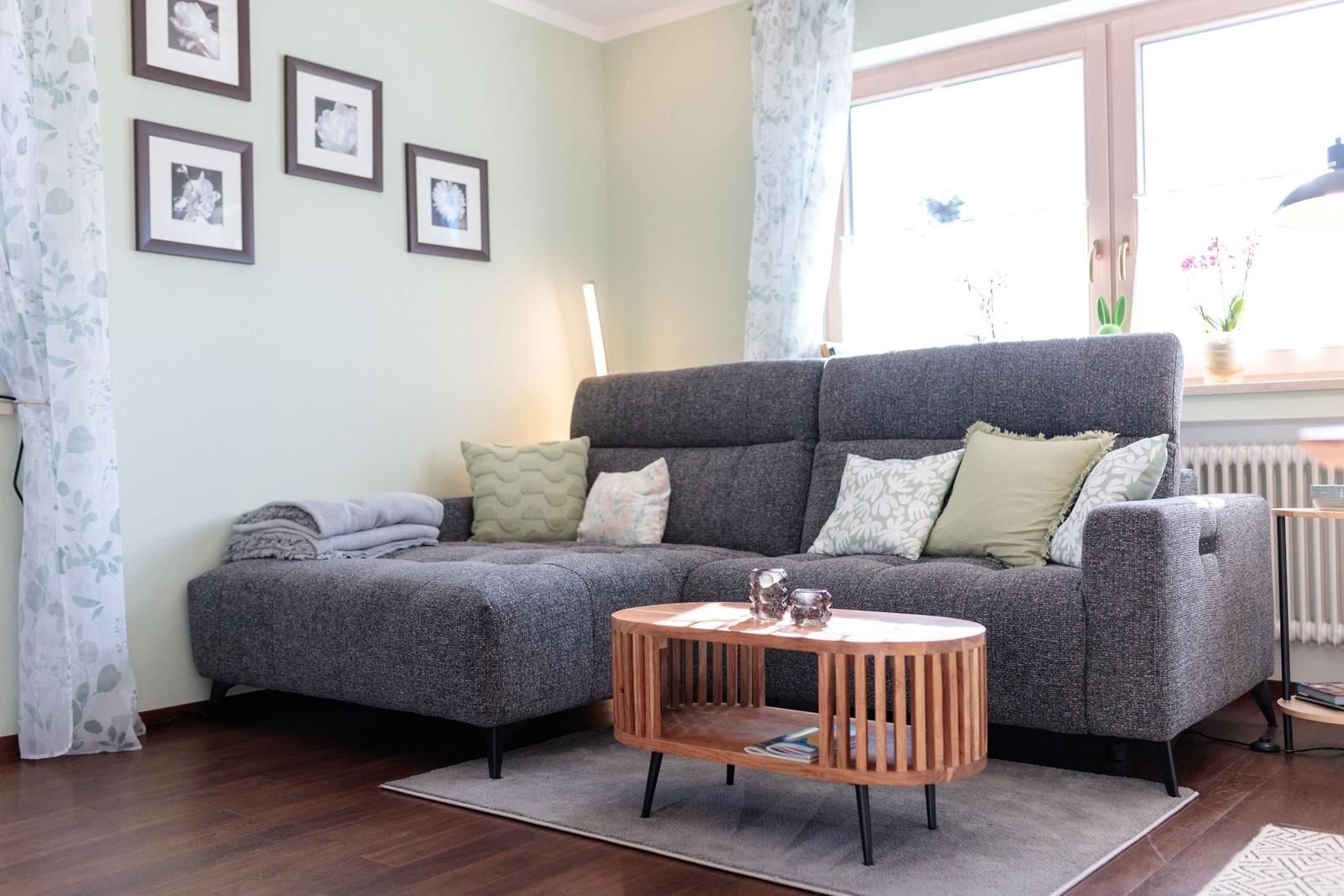 Ein Wohnzimmer mit grauem Sofa, Holztisch und Fenster mit Vorhängen.