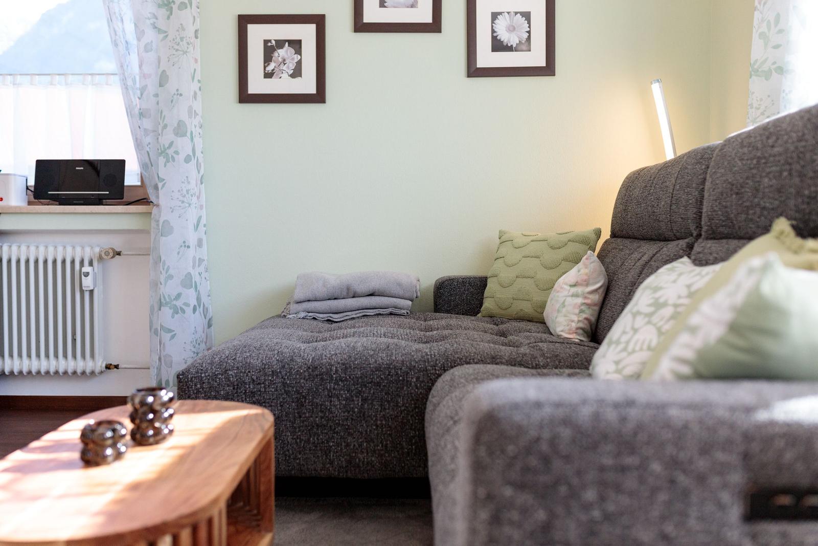 Ein Wohnzimmer mit grauem Sofa, Holztisch und Fenster mit Vorhängen.