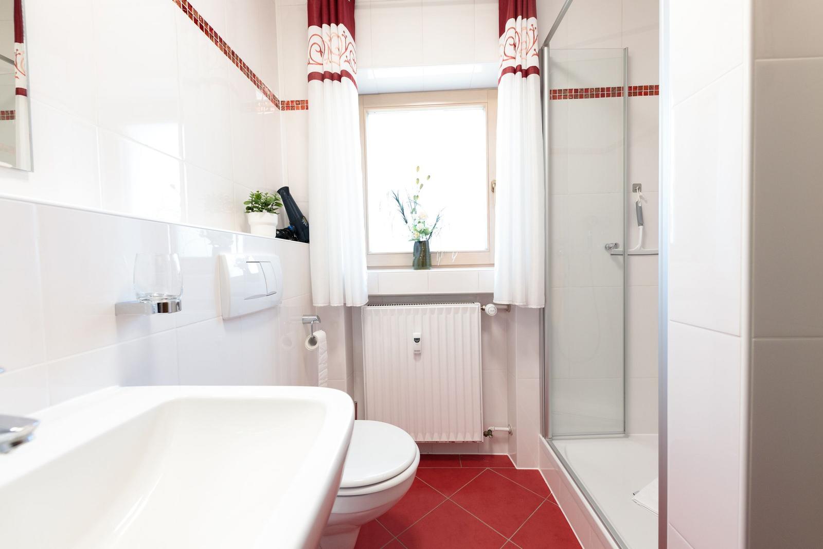 Badezimmer mit Dusche, Toilette und Waschbecken. Rote Fliesen und Fenster mit Vorhängen.