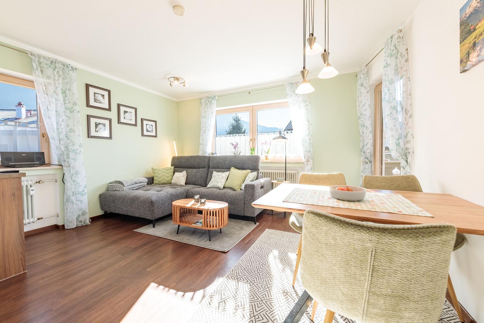 Wohn- und Essbereich mit Sofa, Tisch und Stühlen. Fenster mit Vorhängen und Wandbilder.