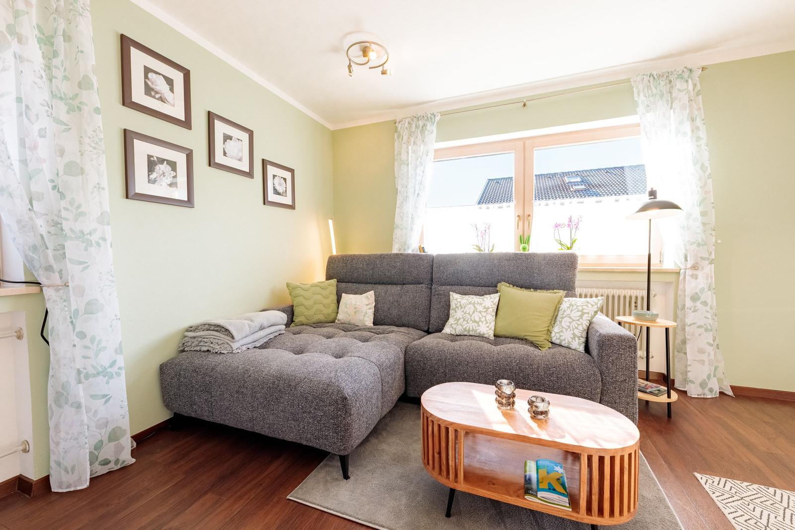 Wohnzimmer mit grauem Sofa, Holztisch und Fenster mit Vorhängen.