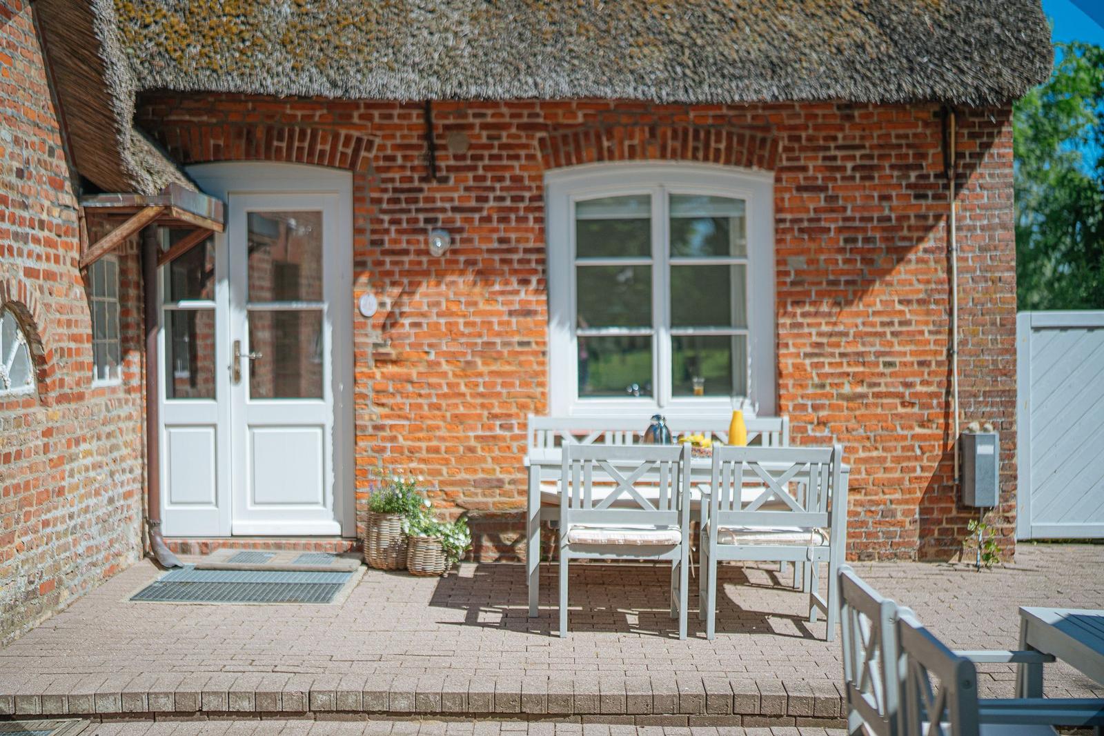 Rotes Backsteinhaus mit Strohdach, weiße Tür und Fenster, Terrasse mit Tisch und Stühlen.