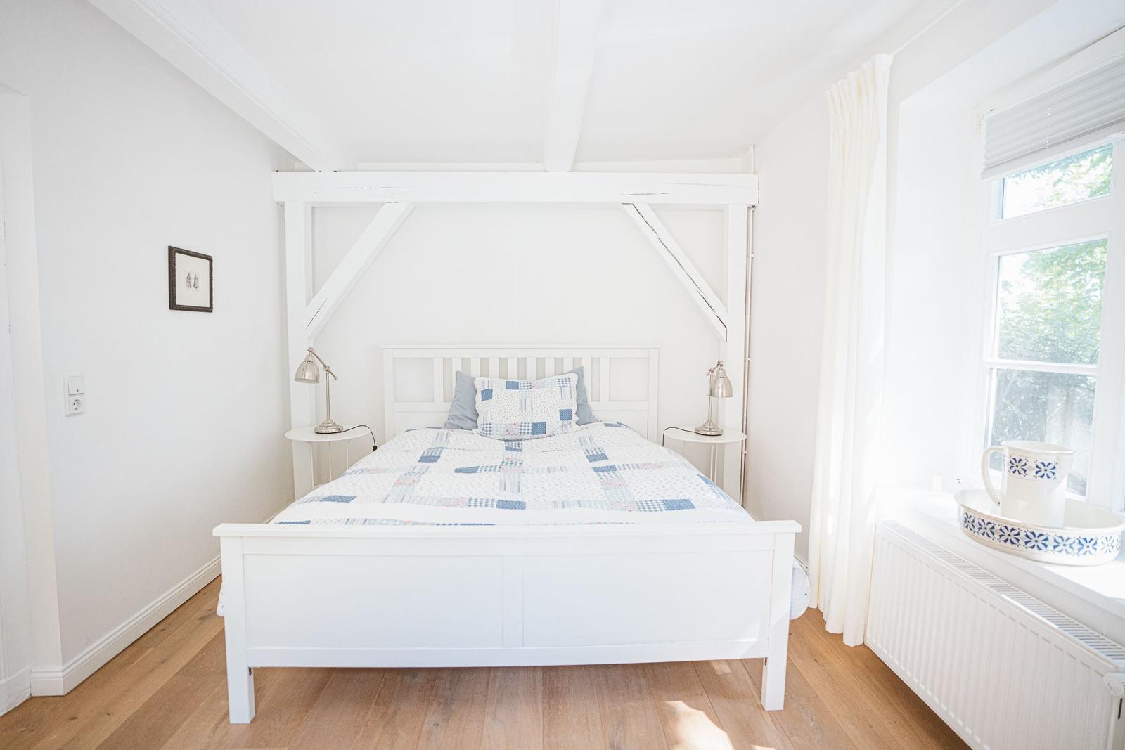 Ein Schlafzimmer mit weißem Bett, Holzboden und Fenster mit Vorhang.