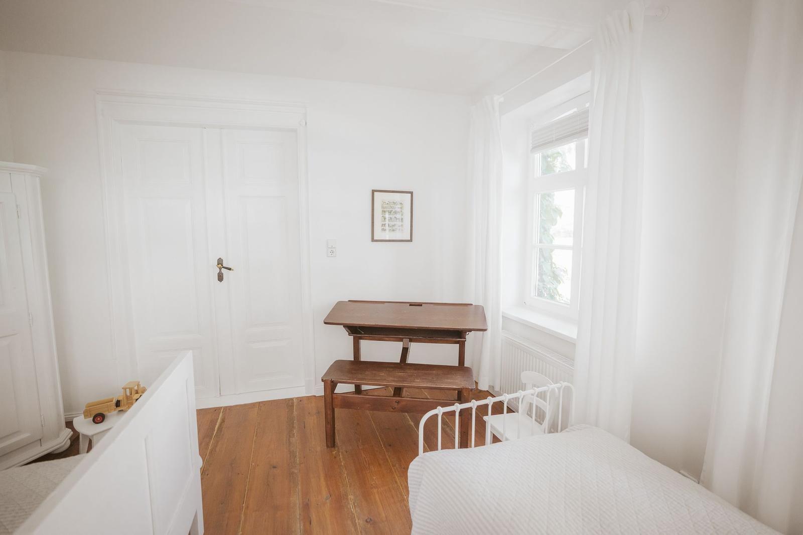 Zimmer mit Holzboden, weißer Tür, Tisch und Bett.