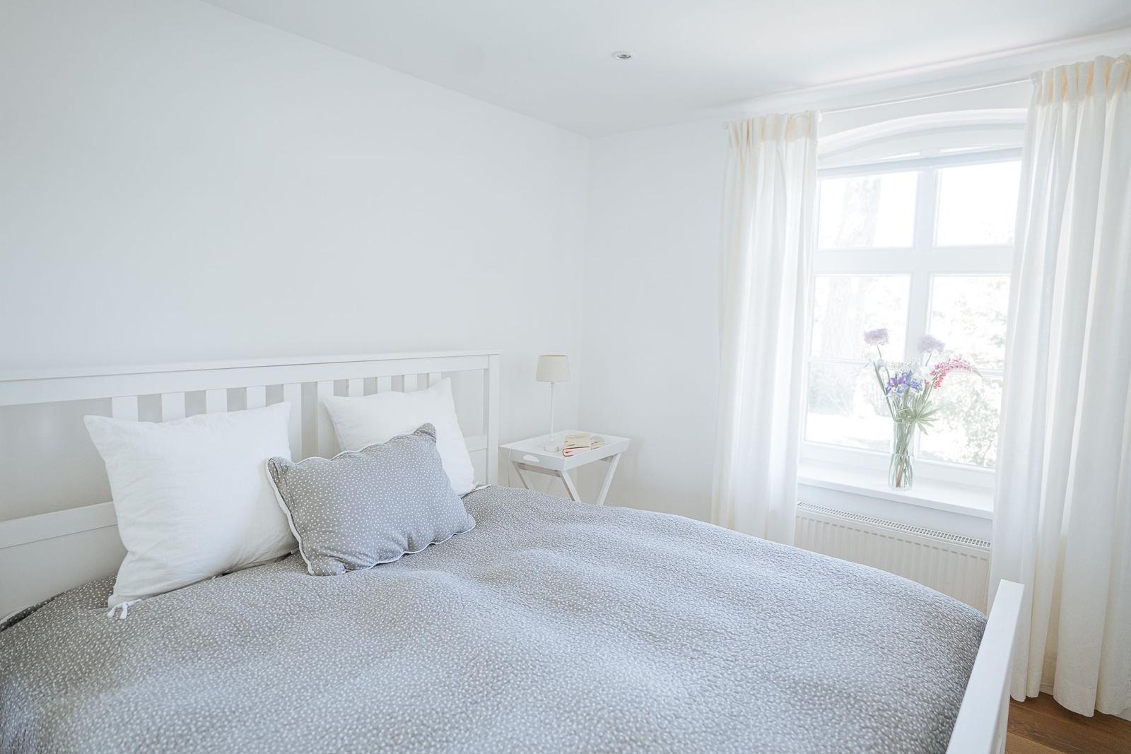 Ein Schlafzimmer mit weißem Bett, Fenster und Blumen auf dem Fensterbrett.