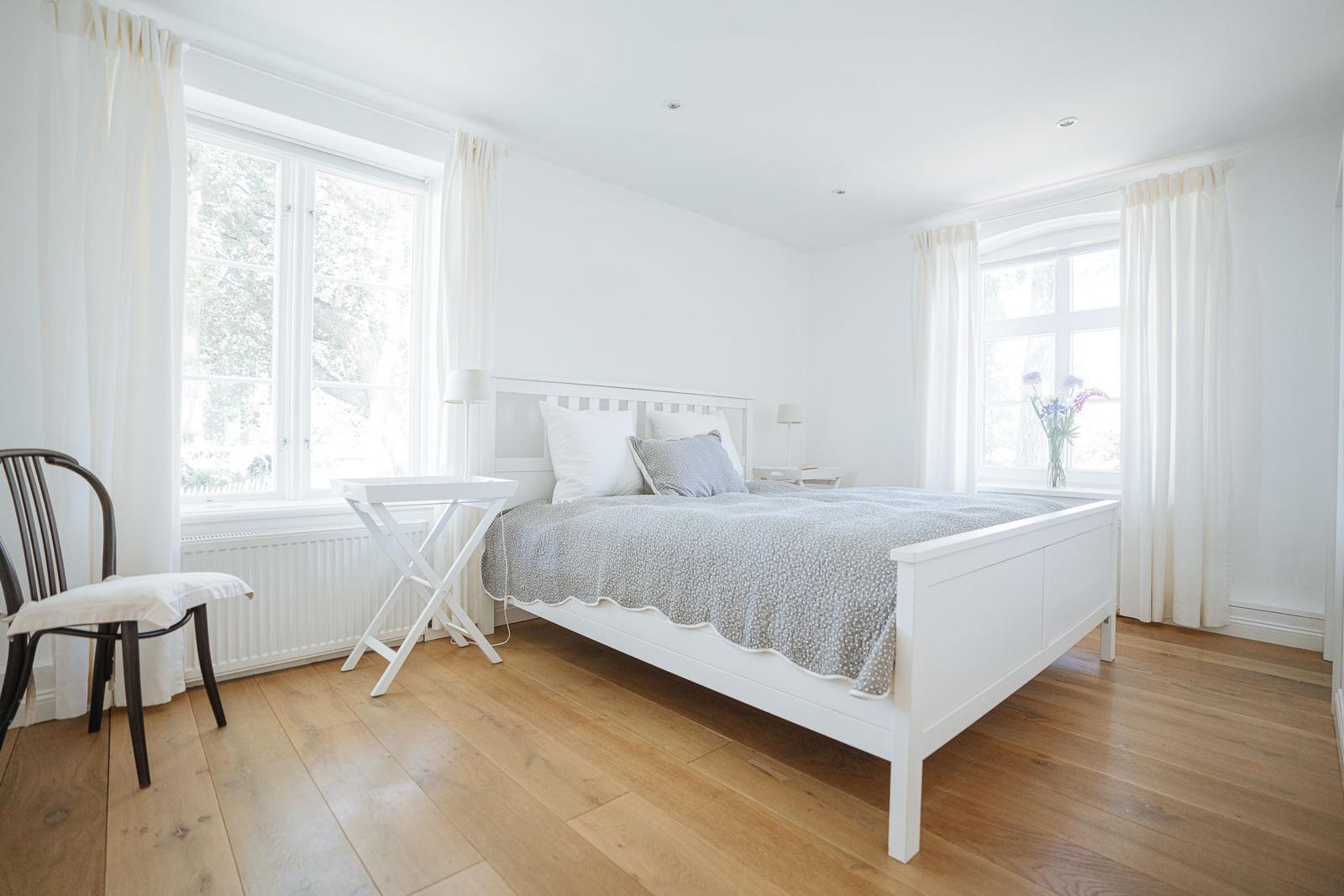 Schlafzimmer mit weißem Bett, Holzboden und zwei Fenstern mit weißen Vorhängen.