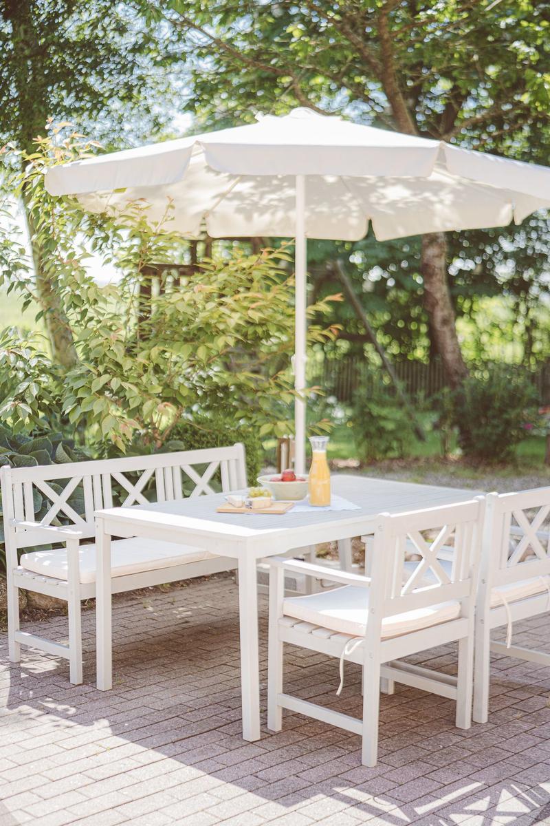 Weiße Terrassenmöbel mit Sonnenschirm auf gepflastertem Bereich unter Bäumen.