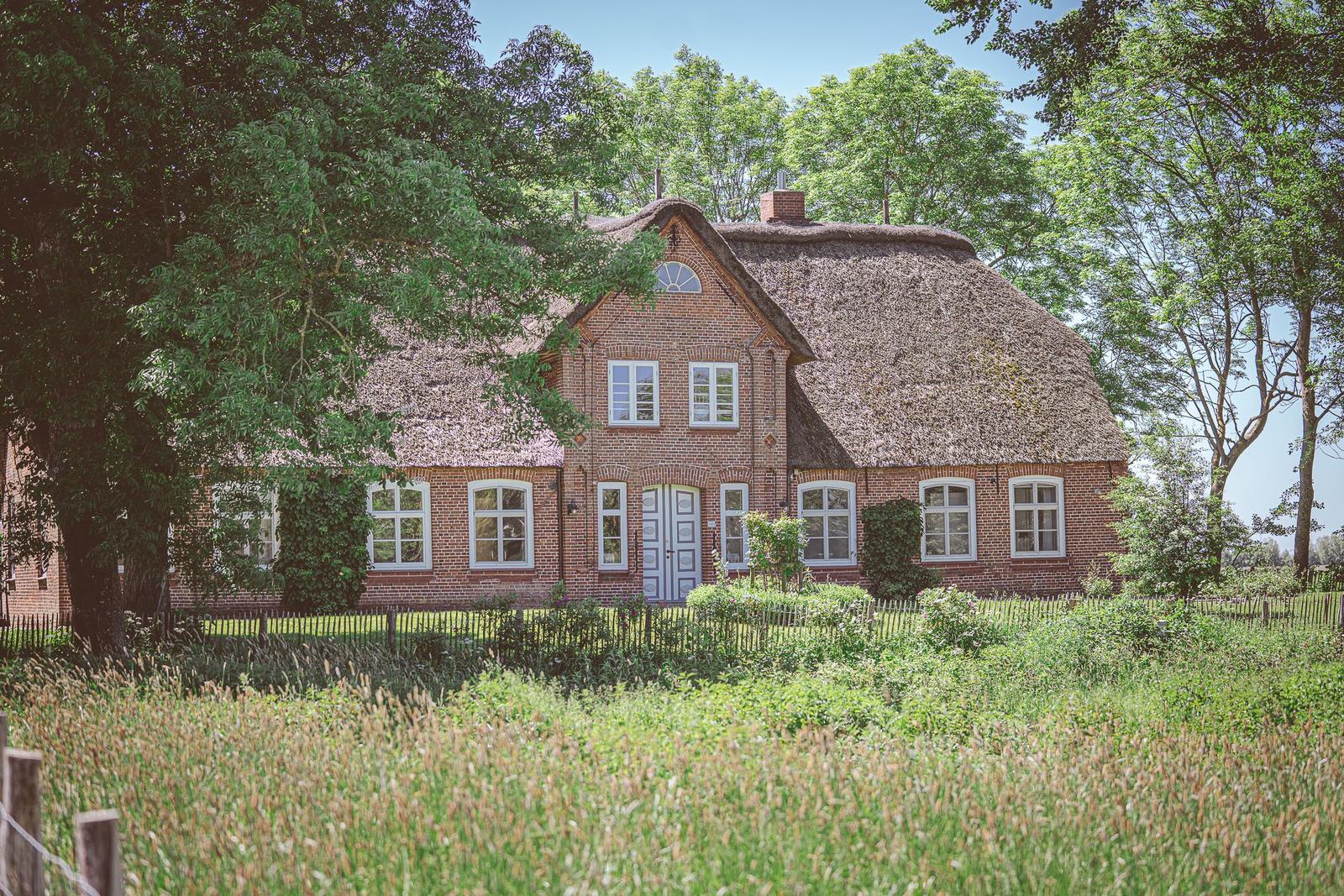 Rotes Backsteinhaus mit strohgedecktem Dach und weißen Fenstern in grüner Umgebung.