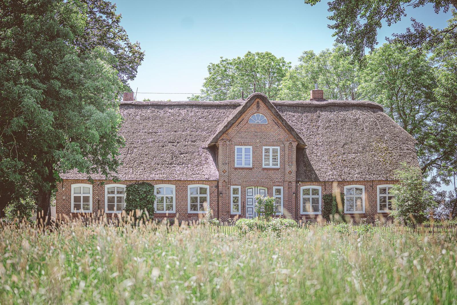 Haus mit strohgedecktem Dach und weißen Fenstern in grüner Umgebung.