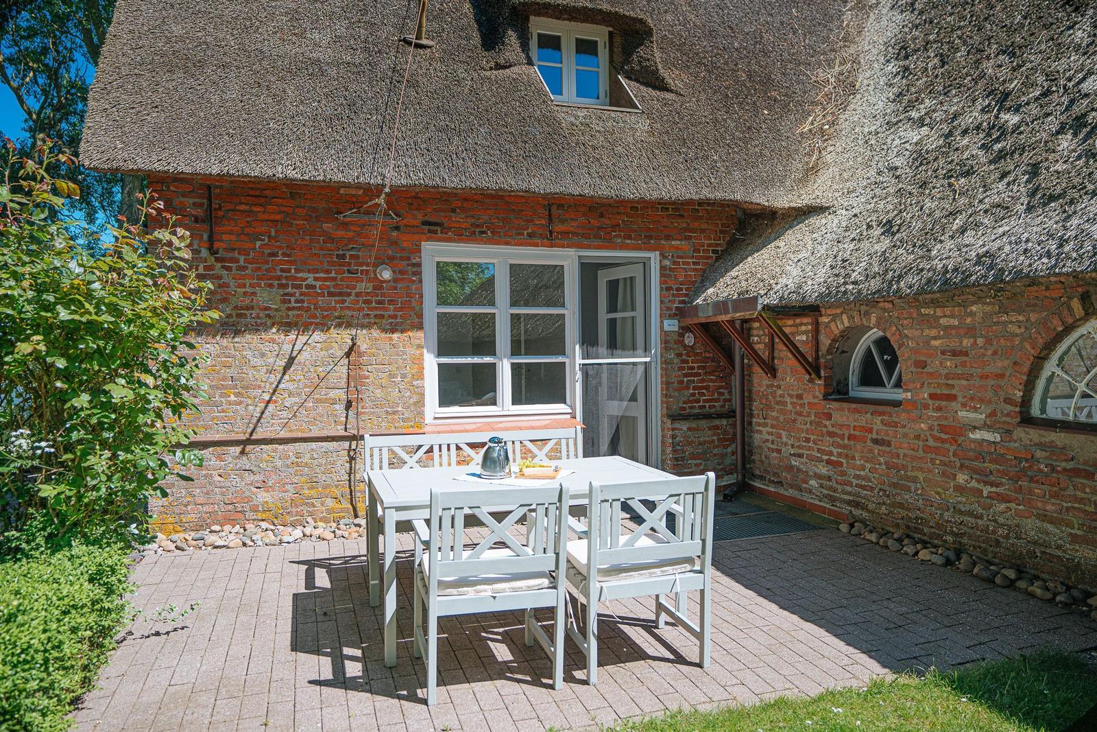 Terrasse mit weißem Tisch und Stühlen vor einem strohgedeckten Haus mit Backsteinmauer.