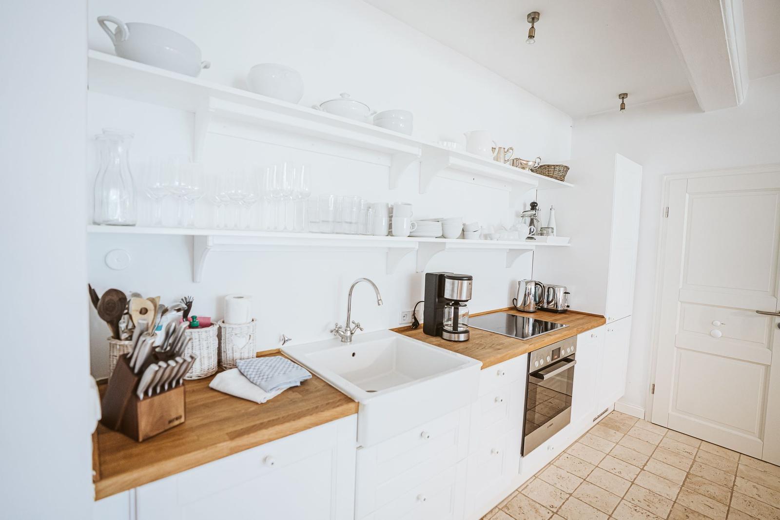 Küche mit Holzplatte, Spüle und weißen Schränken. Auf Regalen stehen Geschirr und Gläser.