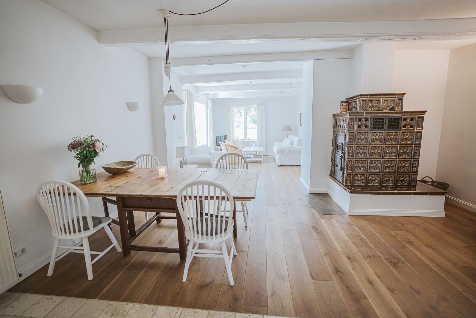 Essbereich mit Holztisch und weißen Stühlen. Im Hintergrund Wohnbereich mit Kamin.